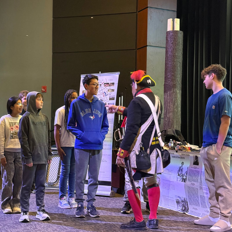 A group of students listens attentively to a person in historical costume at an event.