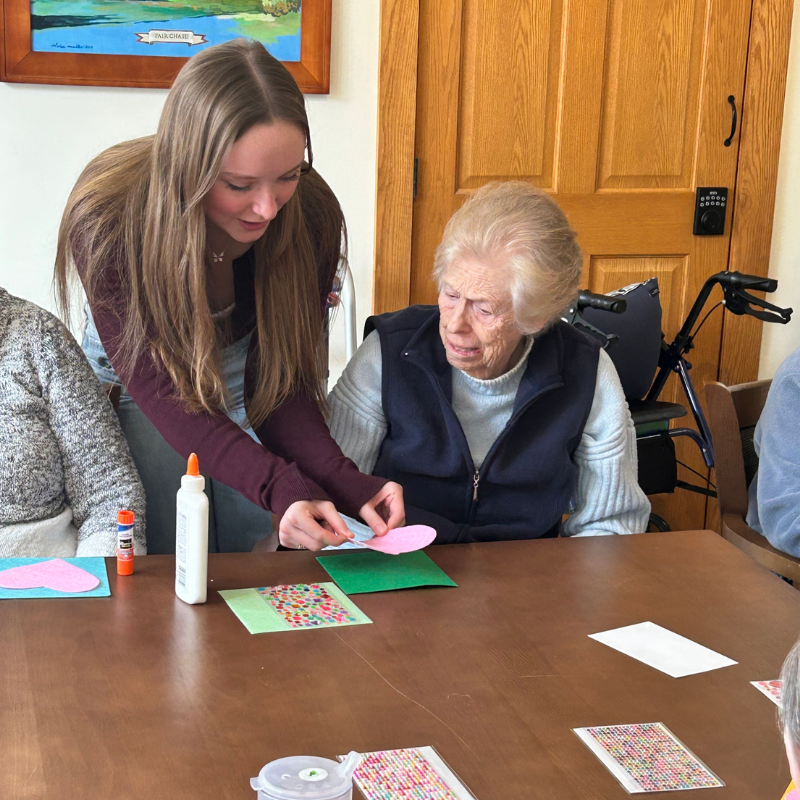 A young woman assists an elderly woman with a craft project at a table.