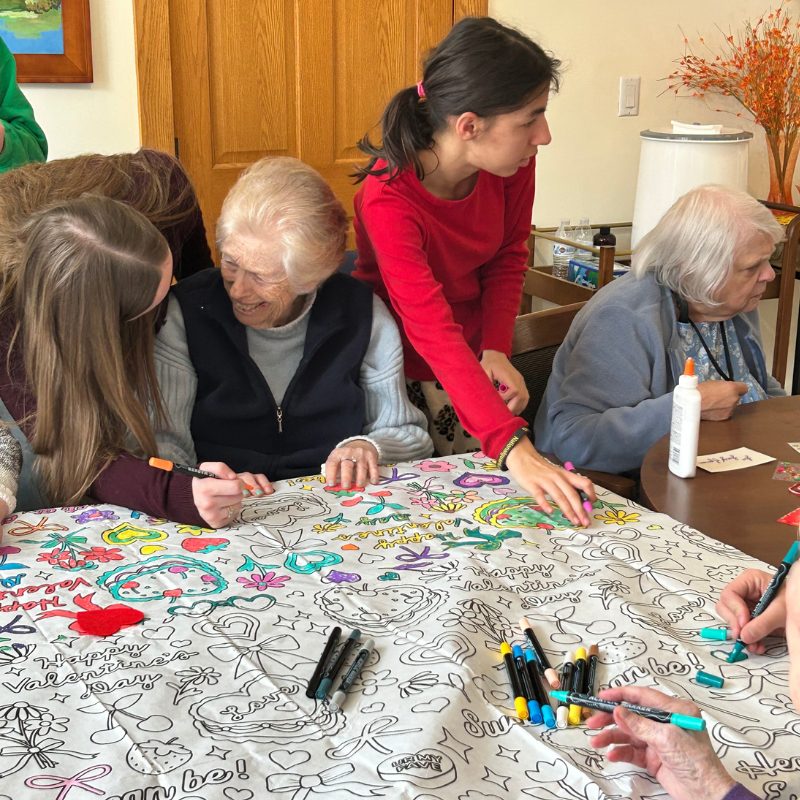 Several people of different ages are coloring a large Valentine's Day themed tablecloth.