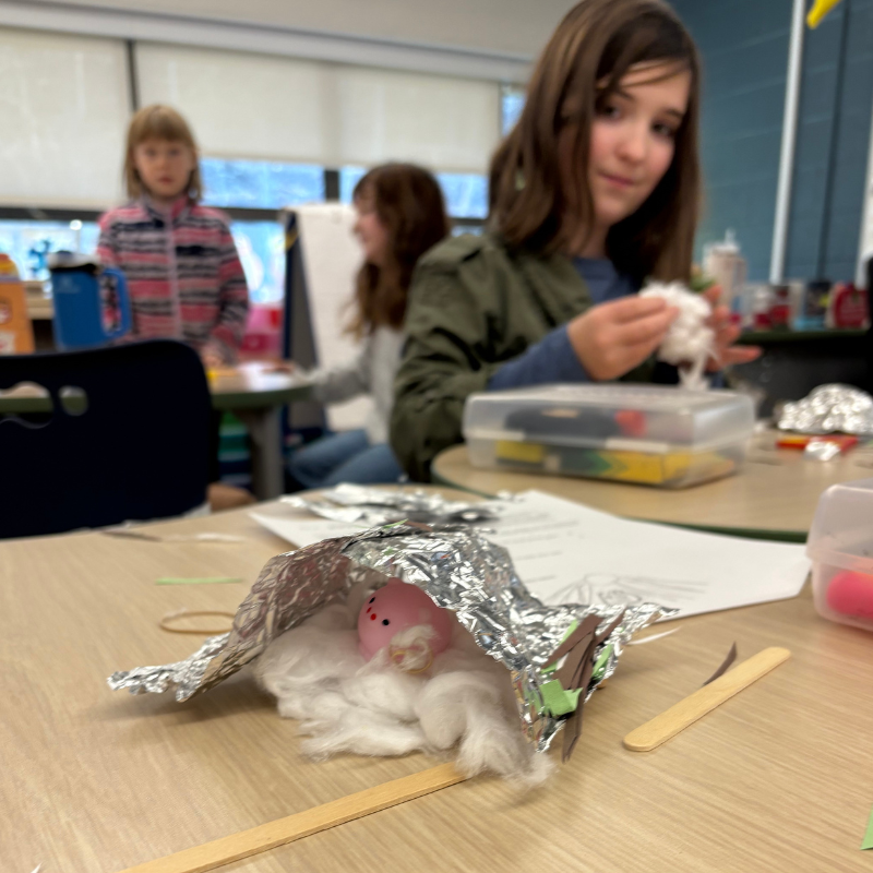 Students work on a craft project at desks in a classroom, with a small pink figure nestled in cotton under foil.
