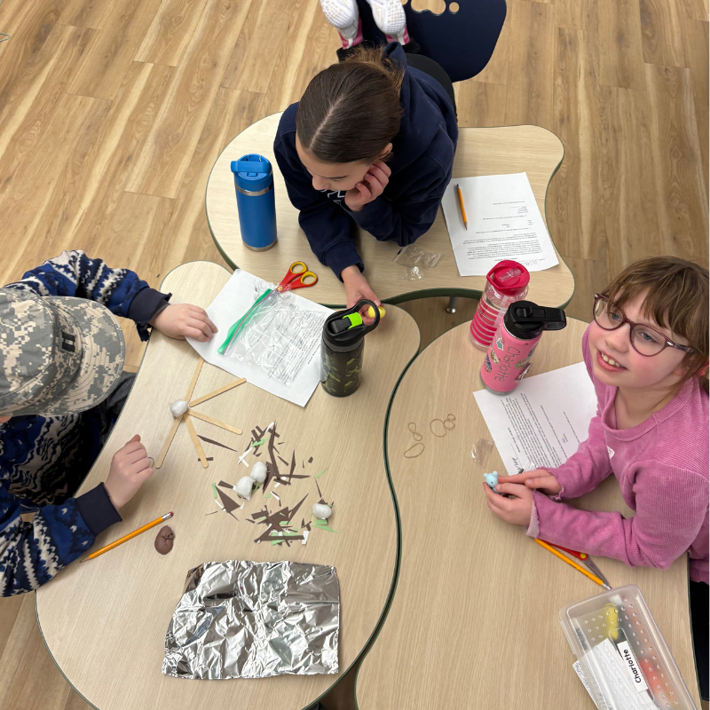 Three children work on a craft project at a table, surrounded by craft supplies like scissors, paper, and cotton balls.