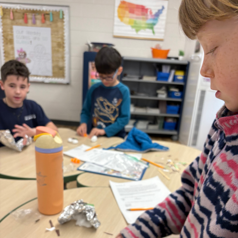 Children work on a craft project at round tables in a classroom, with a map of the US on the wall.