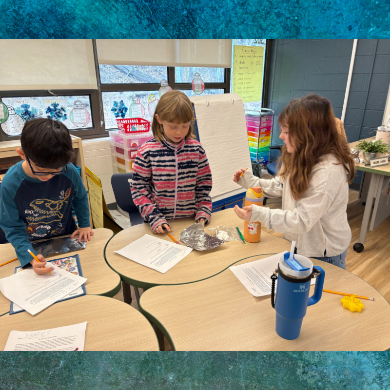 Three children collaborate at round tables in a classroom, working on papers.
