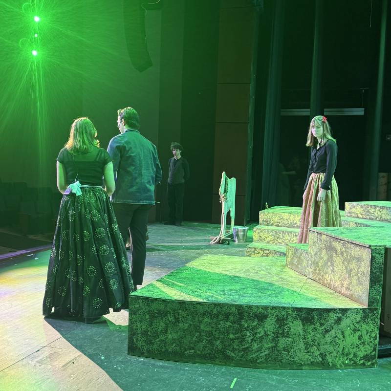 Actors rehearse on a stage with green lighting and tiered platforms.