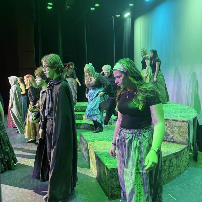 A group of young actors in costume stand on a stage with green lighting.