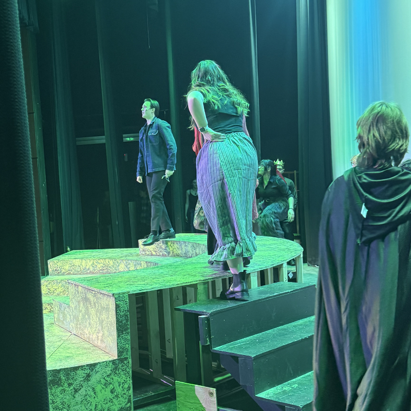 Actors in costume stand on a multi-level stage with green lighting.