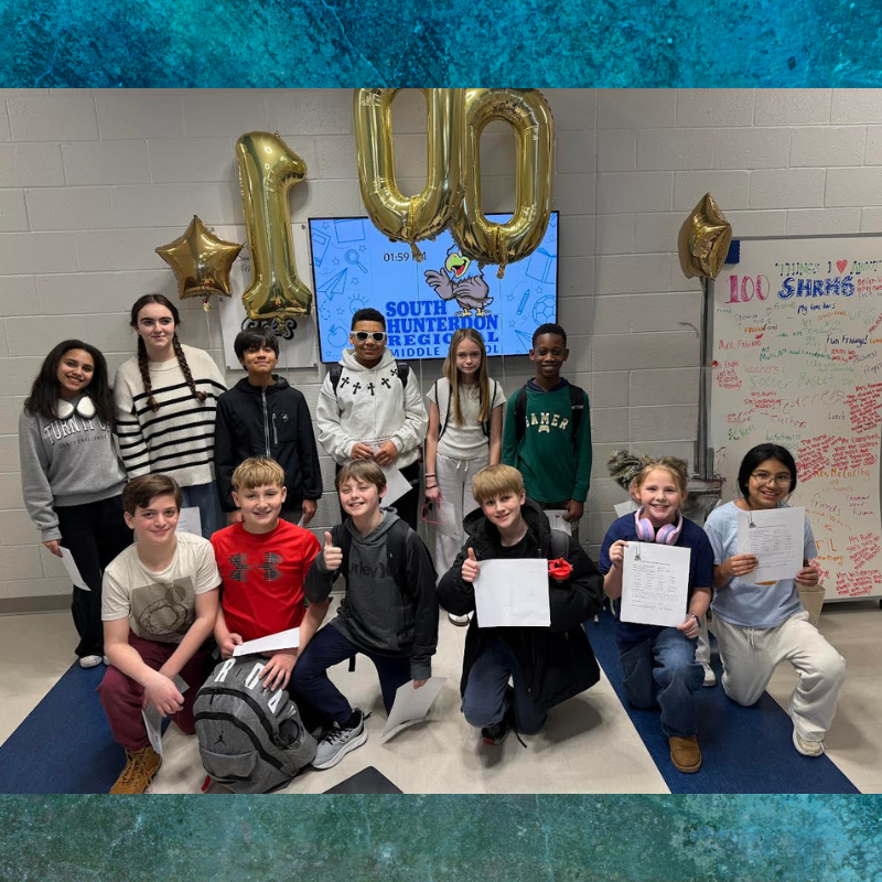 A group of students pose for a photo in front of a screen displaying 'SOUTH HUNTERDON REGIONAL MIDDLE SCHOOL' and large gold balloons spelling '100'.
