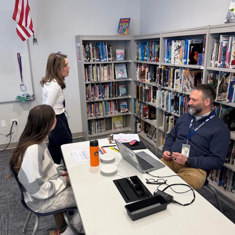A teacher and a student converse in a library, with bookshelves filled with books in the background.
