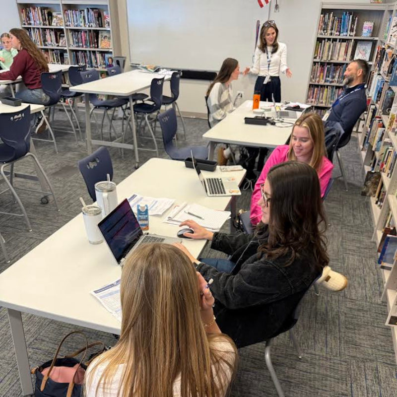 Students work collaboratively on laptops at tables in a library setting with bookshelves in the background.