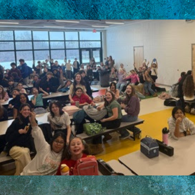 A large group of students are gathered in a school cafeteria, many smiling and looking towards the camera.