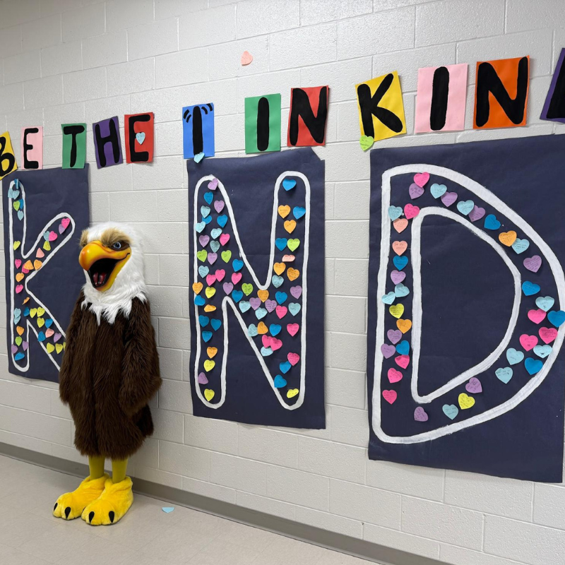 A mascot in an eagle costume stands next to large letters spelling 'KIND', decorated with colorful paper hearts.