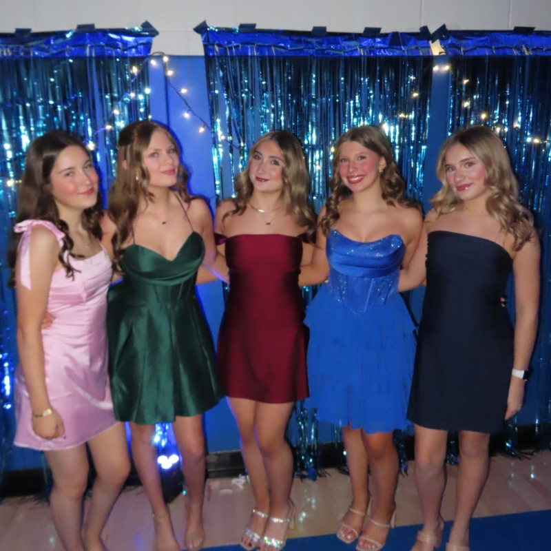 Five young women in elegant dresses pose for a photo in front of a blue sequined backdrop.