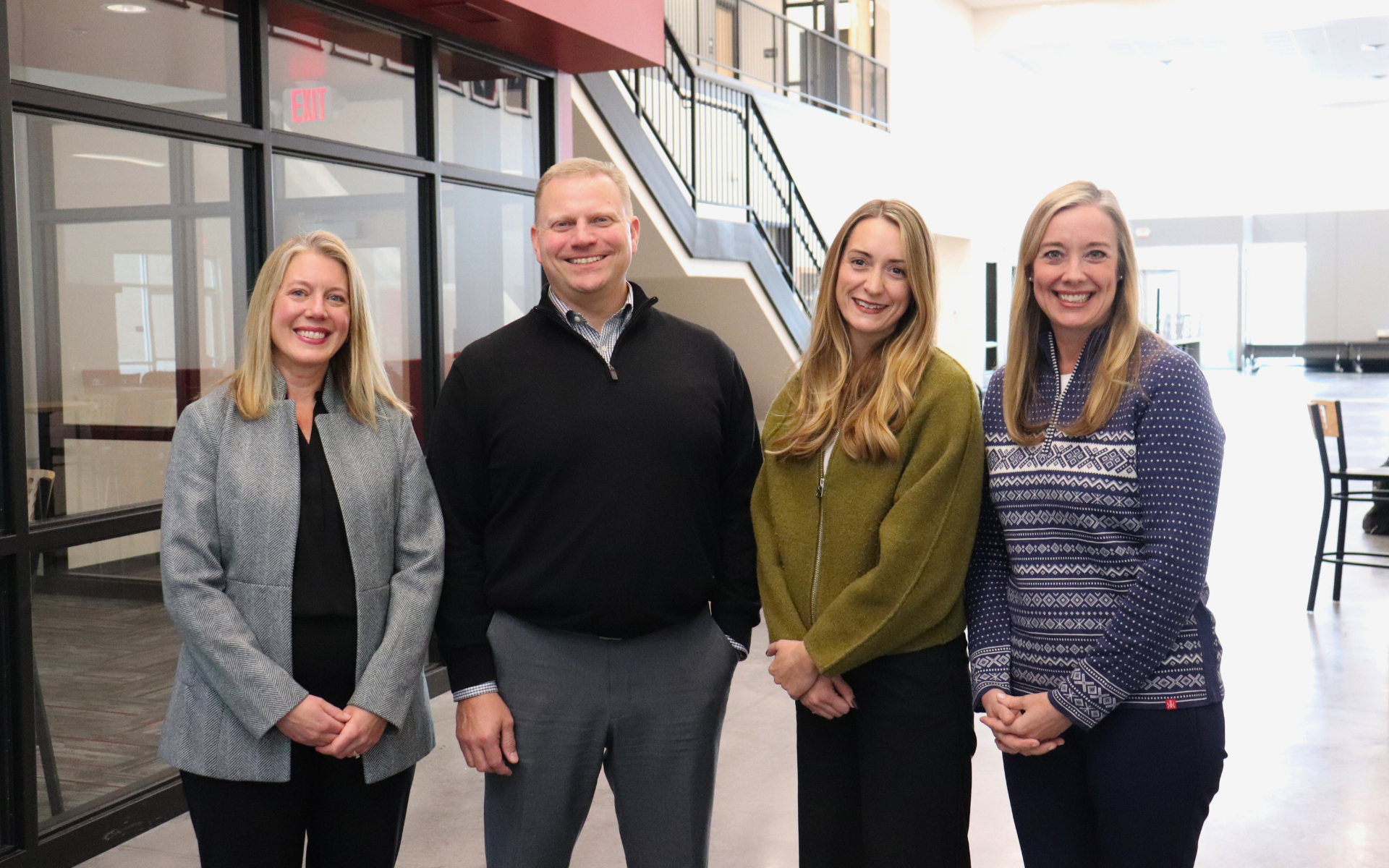 Four smiling individuals stand together in a modern office hallway with a staircase in the background.