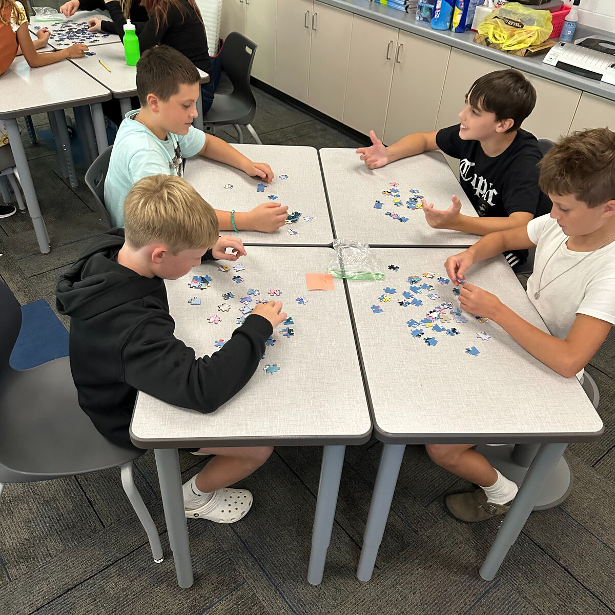 Four children sit at connected desks in a classroom, engaged in putting together jigsaw puzzles.