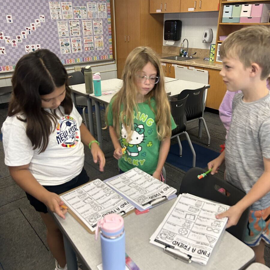Three children stand around school desks, reviewing worksheets titled "FIND A FRIEND" in a brightly lit classroom.