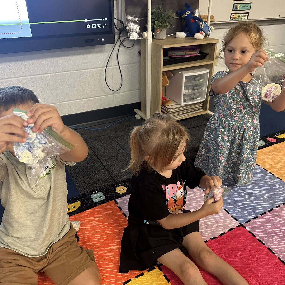 Three children sit on a colorful classroom rug, holding up clear plastic bags with art projects inside.