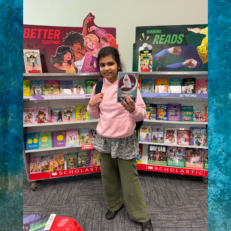 A young person holds up a book in front of a colorful display of children's books.