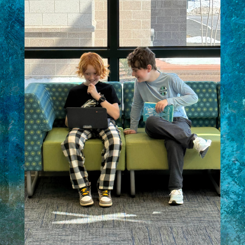 Two boys sit on a green couch, one using a laptop and the other holding a book.