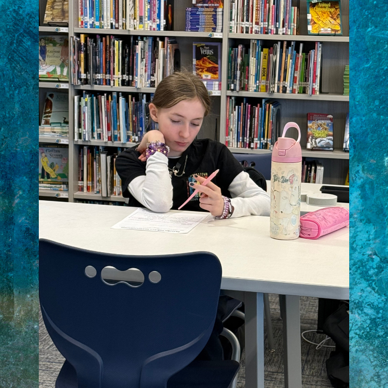 A young person studies at a table in a library, surrounded by bookshelves.