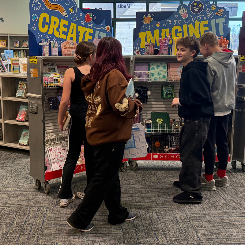Four young people browse a display of art and school supplies with "CREATE IT" and "IMAGINE IT" signs.