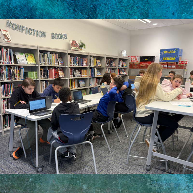 Students work on laptops at tables in a library with shelves of books.