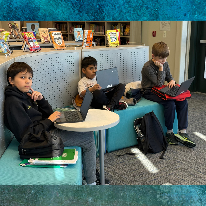 Three boys focus on their laptops in a library setting with bookshelves in the background.