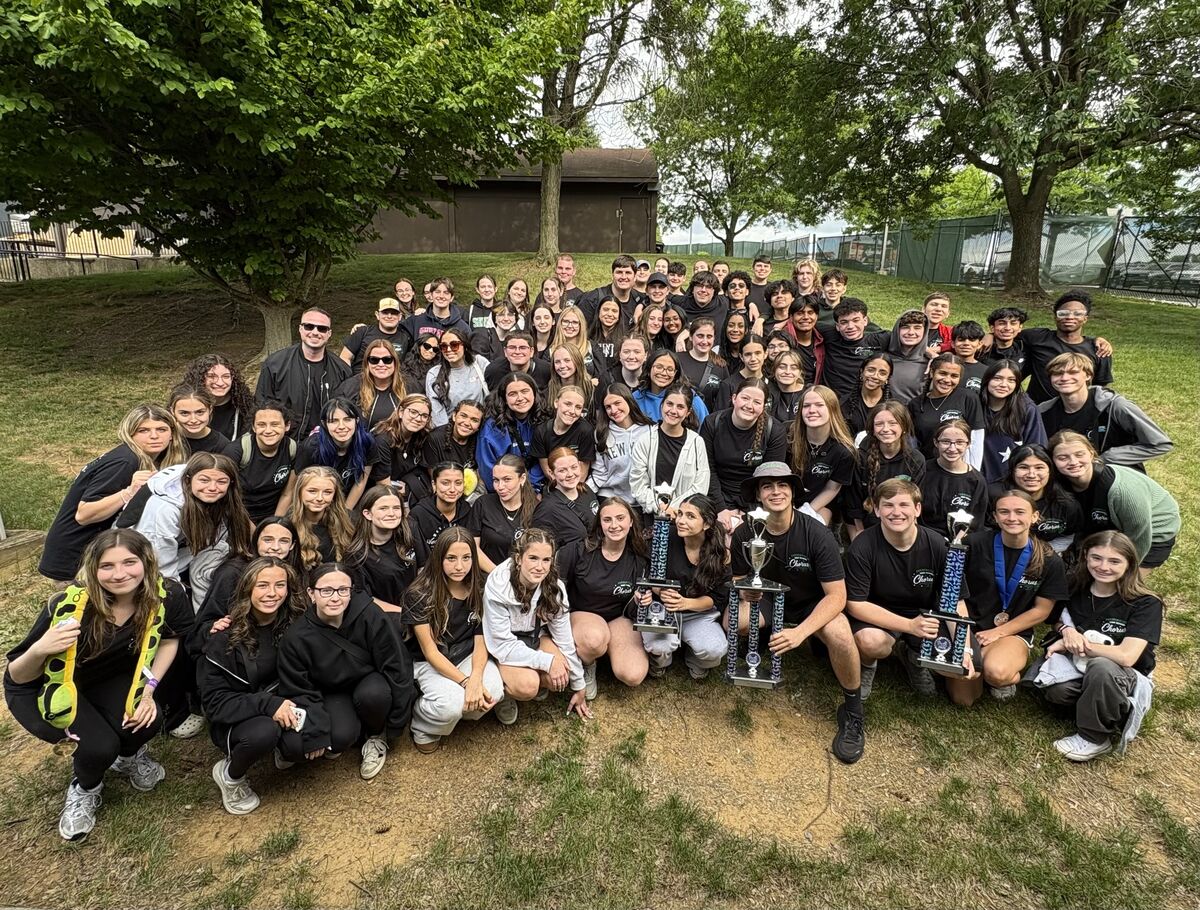 A large group of students in black t-shirts pose for a photo outdoors, some holding trophies.