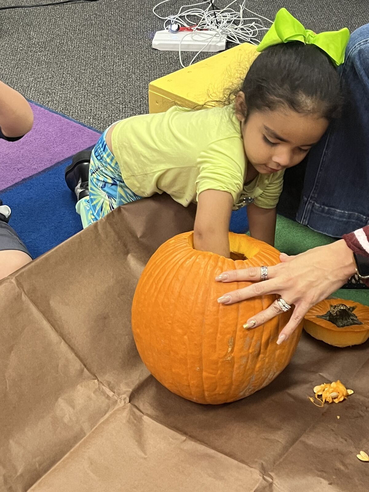A young child reaches inside a large orange pumpkin while an adult guides the process.