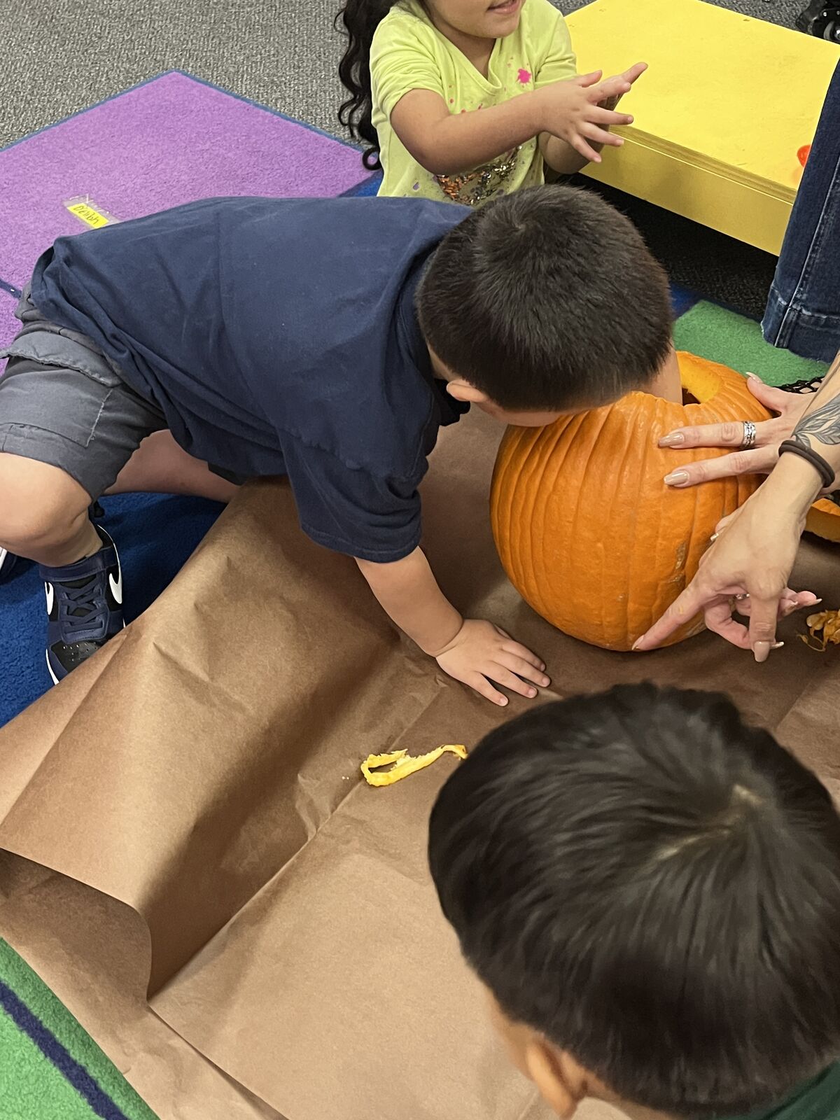 An adult guides a child's hands to carve a large orange pumpkin on a brown paper-covered table.