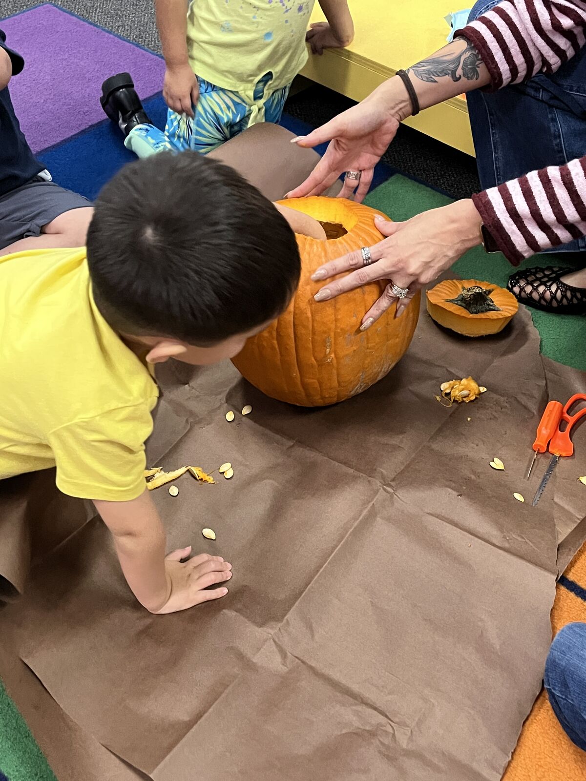 A child in a yellow shirt helps carve a large orange pumpkin on brown paper.
