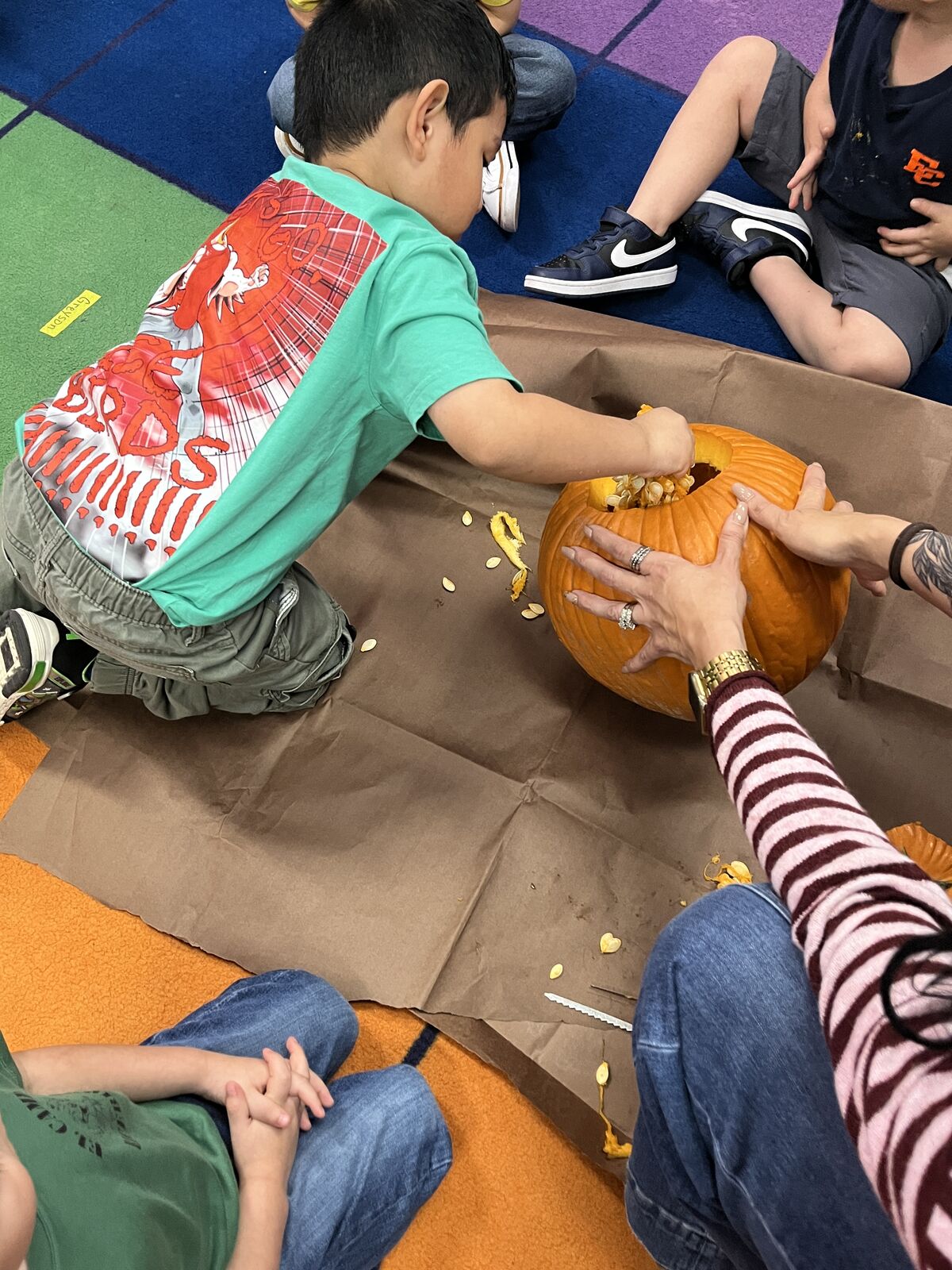 A child in a green shirt reaches inside a pumpkin to scoop out seeds on a brown paper-covered floor.