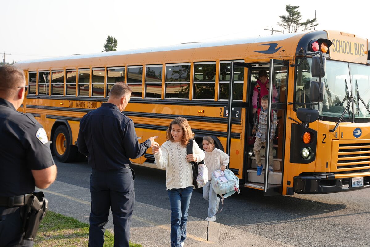 A first responder in uniform hands a small item to a young girl getting off a yellow school bus.