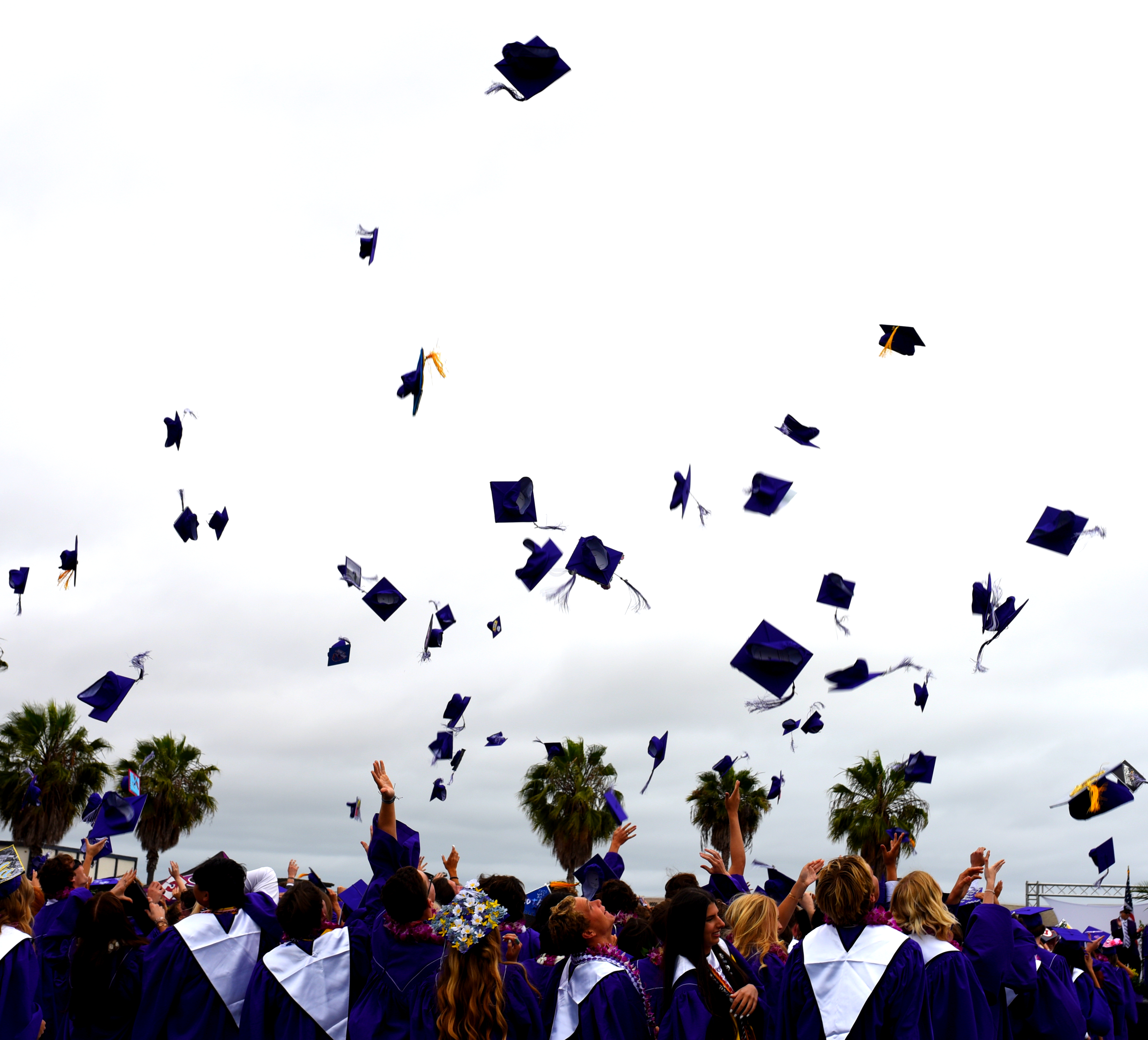 Graduates in purple gowns toss their caps in the air under a cloudy sky.