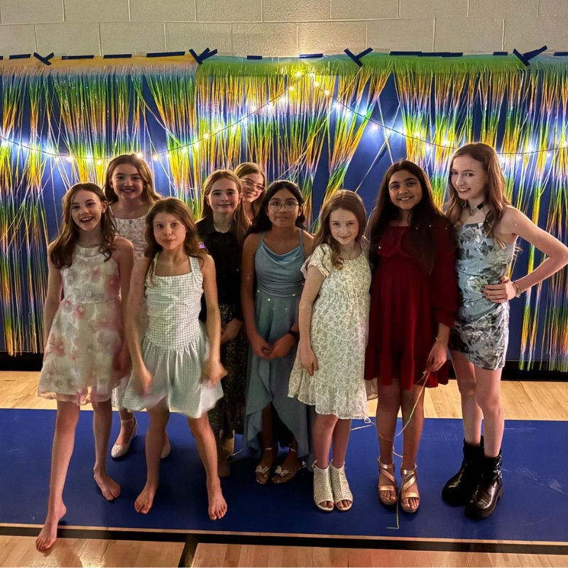 A group of young girls in dresses pose for a photo in front of a colorful, shimmering backdrop.