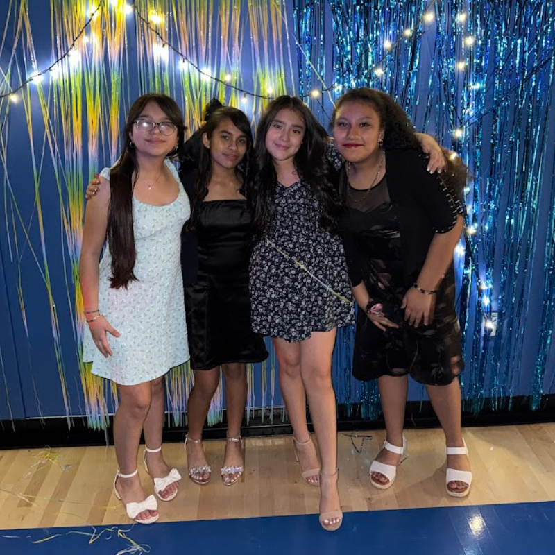 Four young women pose for a photo in front of a blue sequined backdrop with string lights.