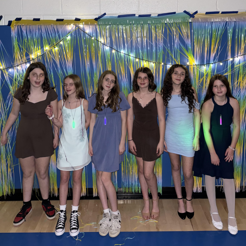 Six young women in dresses pose for a photo in front of a blue and yellow fringe backdrop with string lights.