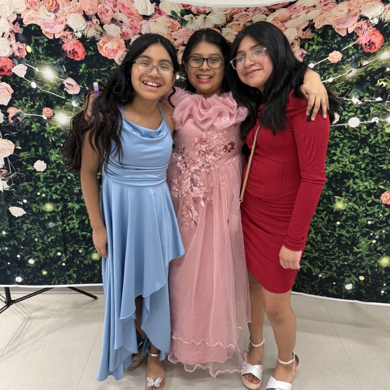 Three smiling young women pose for a photo in front of a floral backdrop.