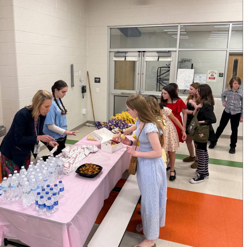 A group of young people and an adult stand around a table laden with snacks and bottled water.