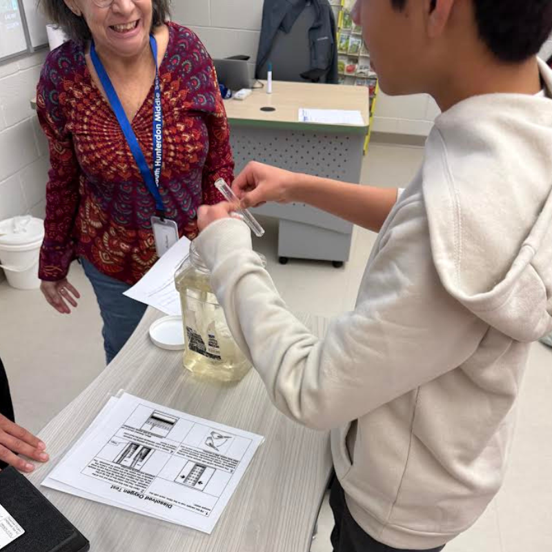 A teacher smiles as a student performs a dissolved oxygen test in a classroom.