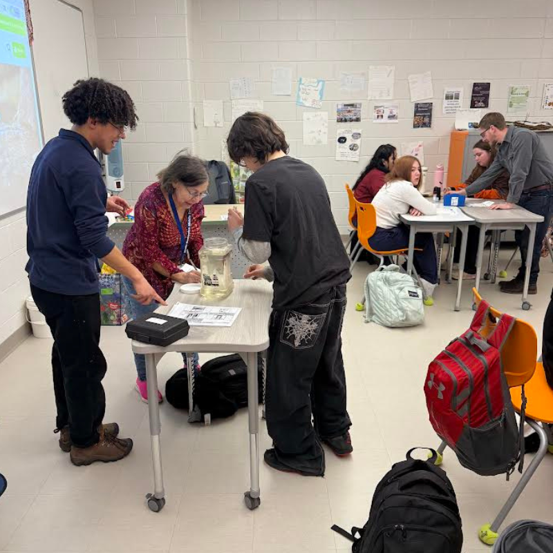 Students and a teacher gather around a table in a classroom, observing a jar of liquid.