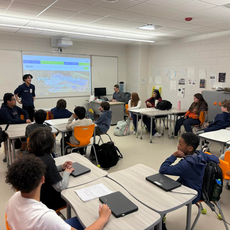 A young presenter stands by a projector screen displaying "Visit our Center & Reserve" to a classroom of students.