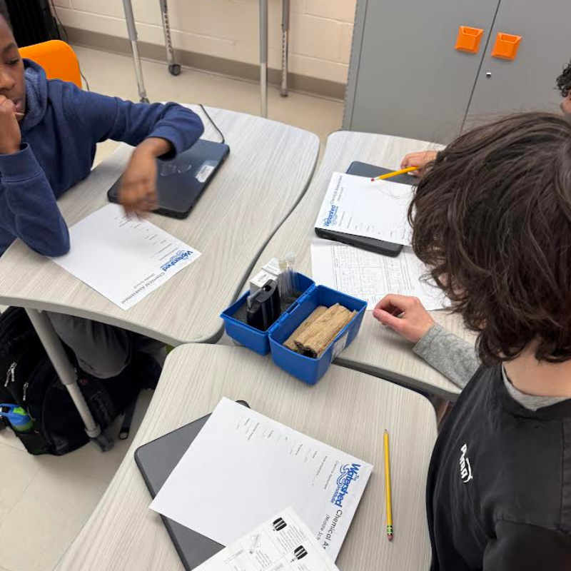 Students work at desks with science worksheets and lab equipment, including test tubes and wooden samples.
