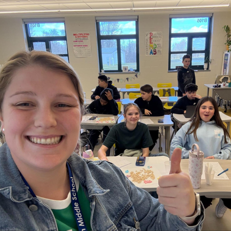 A smiling teacher takes a selfie in a classroom with students at their desks.