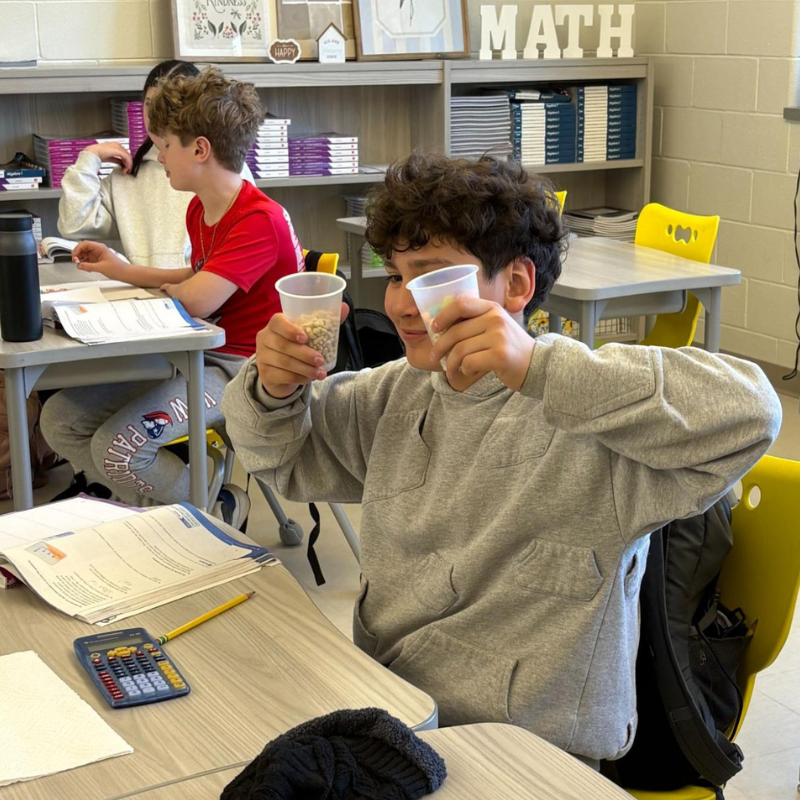 A young boy in a classroom holds up two clear cups, one filled with small, light-colored objects.