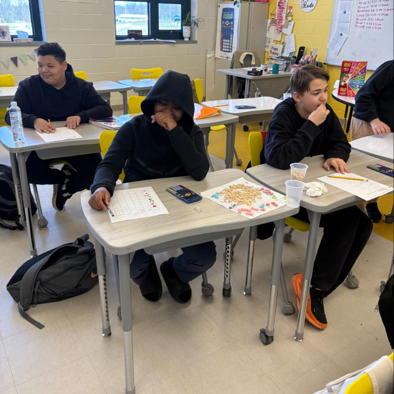 Students work at desks in a classroom, with papers and small colorful objects on their tables.