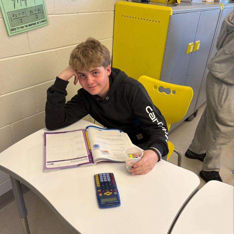 A young person sits at a desk with a math textbook and calculator, holding a cup of colorful candies.