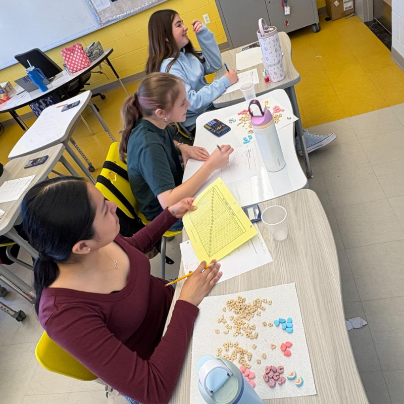 Three students work at desks in a classroom, with papers and colorful cereal scattered on one desk.