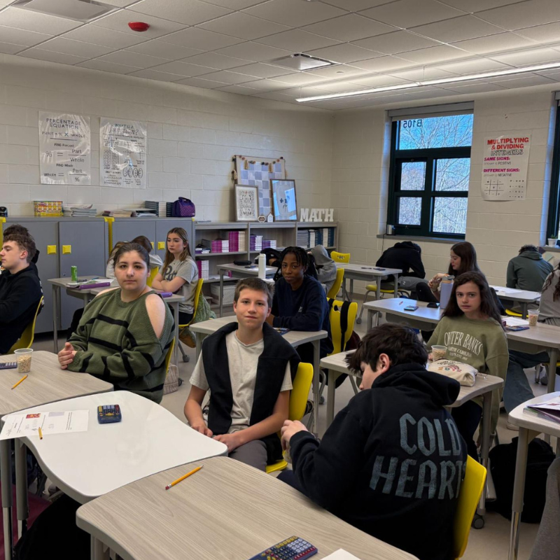 Students sit at desks in a classroom, facing forward, with math posters on the wall.