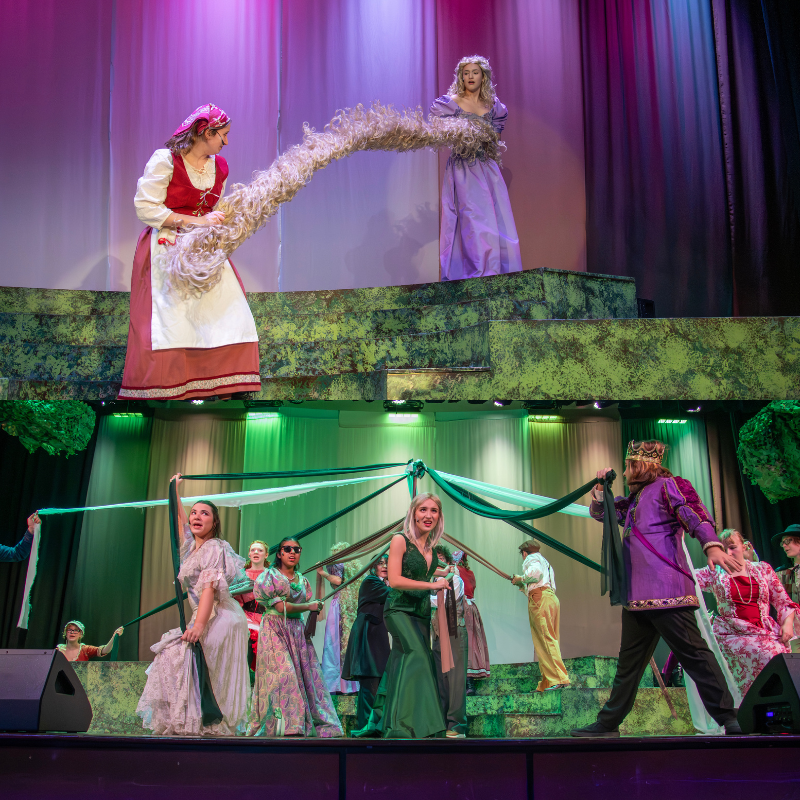 A theatrical scene with characters on a multi-level stage, one holding a long blonde wig.