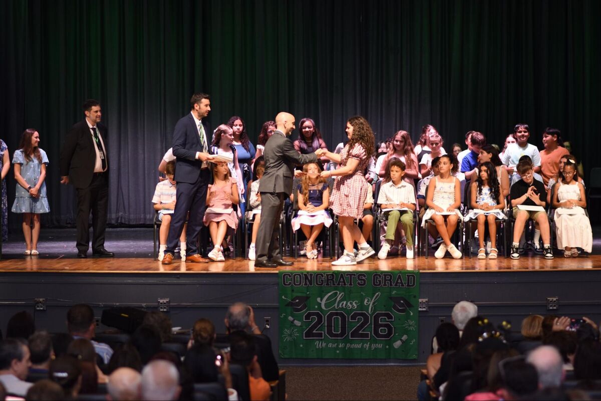 Students in formal attire are recognized on stage during a graduation ceremony, with a banner reading 'CONGRATS GRAD Class of 2026'.